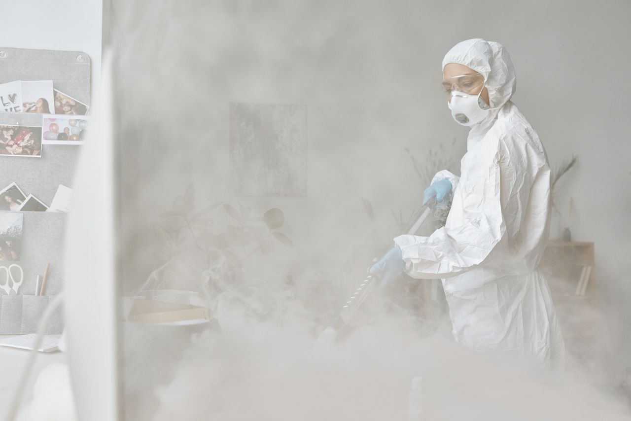 A woman wearing protective gear disinfects an indoor area using fogging equipment.